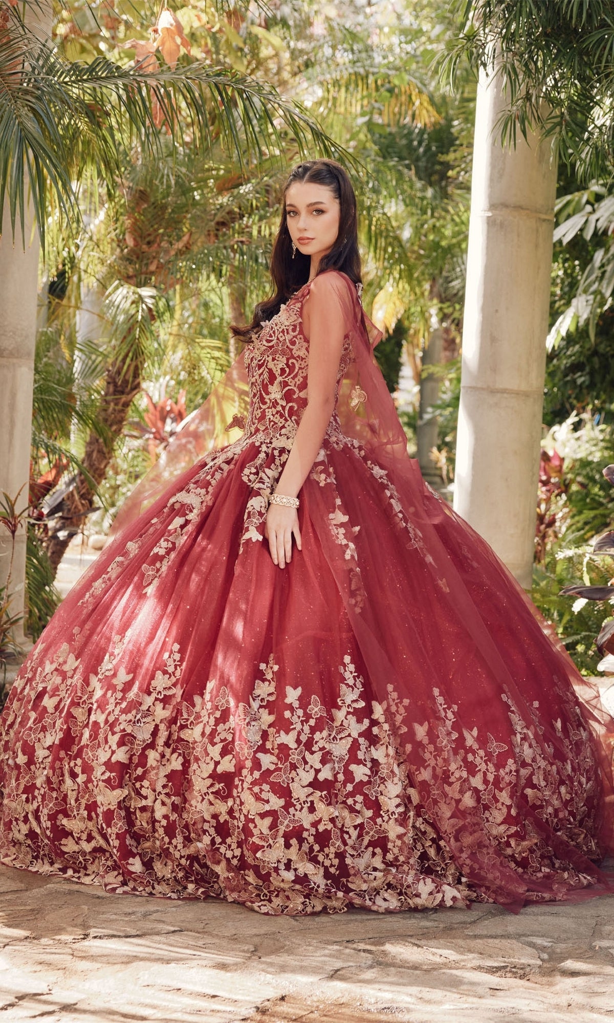 A woman stands outdoors in a garden wearing the Juliet 1442 Quinceañera Dress with Sheer Cape, featuring a voluminous burgundy skirt and gold floral embroidery, looking over her shoulder.